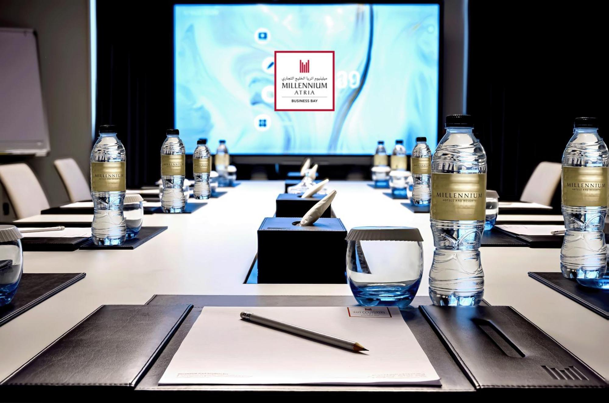 Aparthotel Millennium Atria Business Bay Dubai Exterior foto The photo shows a modern conference room setup. A long table is laid out with neatly arranged stationery, including notebooks and pens, at each seat. Several bottles of water are placed on the table. In the background, a large screen is displayed, li