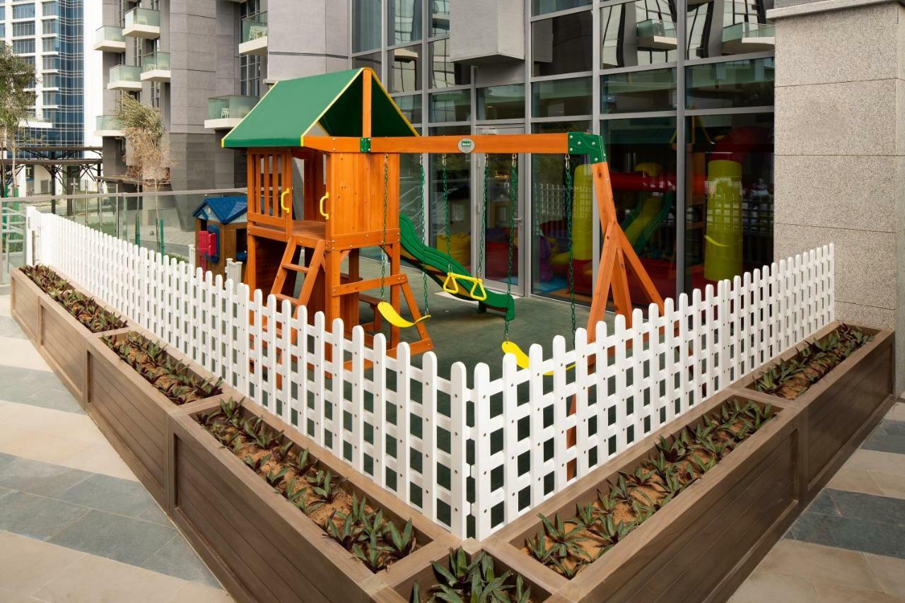 Aparthotel Millennium Atria Business Bay Dubai Exterior foto The image shows a children's playground area, which is enclosed by a white picket fence. The playground features a wooden play structure with a slide, swings, and climbing elements. Surrounding the play area are planters with greenery, and the backdr