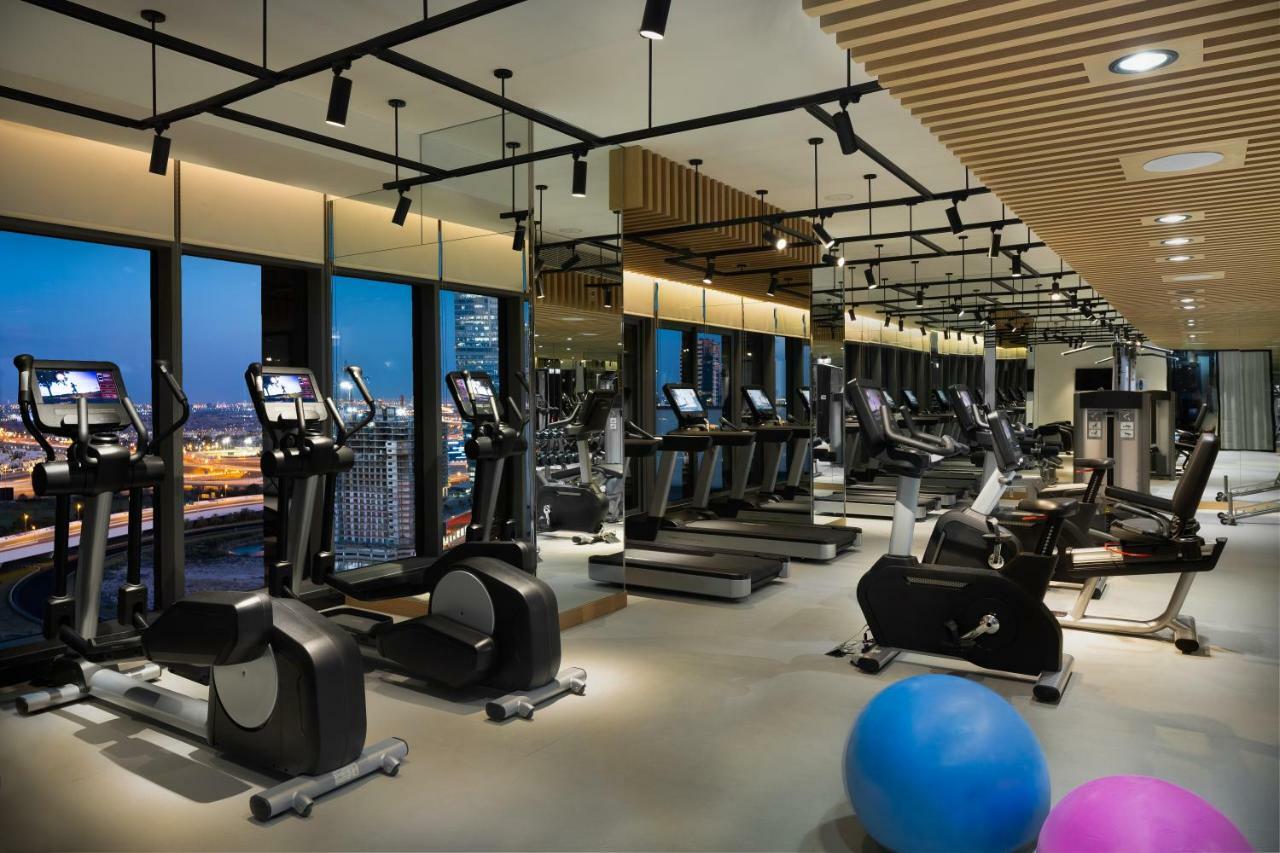 Aparthotel Millennium Atria Business Bay Dubai Exterior foto The photo shows a modern gym with various types of exercise equipment available. There are several treadmills and elliptical machines arranged in rows, with large windows in the background providing a view of the city. The lighting is bright and cont
