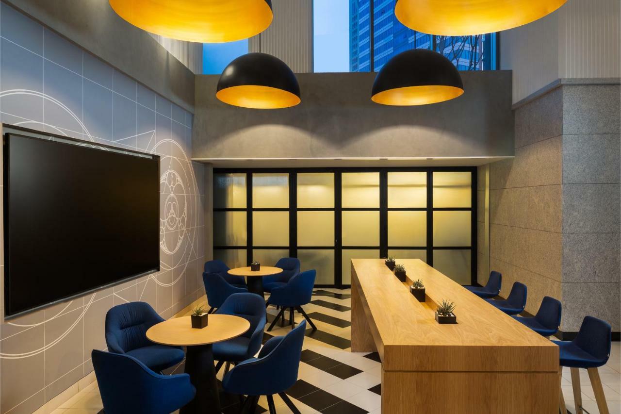 Aparthotel Millennium Atria Business Bay Dubai Exterior foto The photo shows a modern interior space, likely a lounge or meeting area. It features a long wooden table with some small plants on it, accompanied by several blue, circular chairs arranged around it. There is a large flat-screen television mounted o